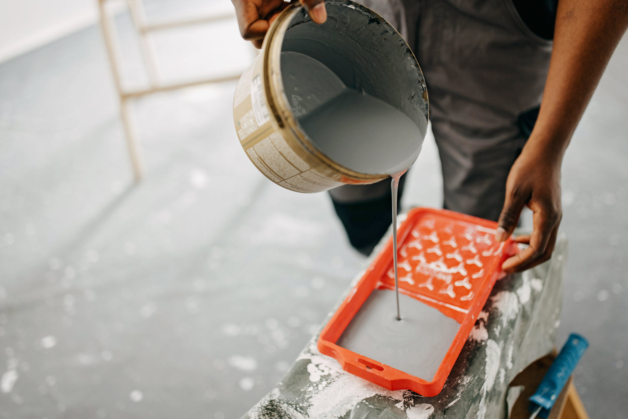 Afroamerikansk målare i arbetsoverall häller vit färg från hink till orange målarbricka inomhus
