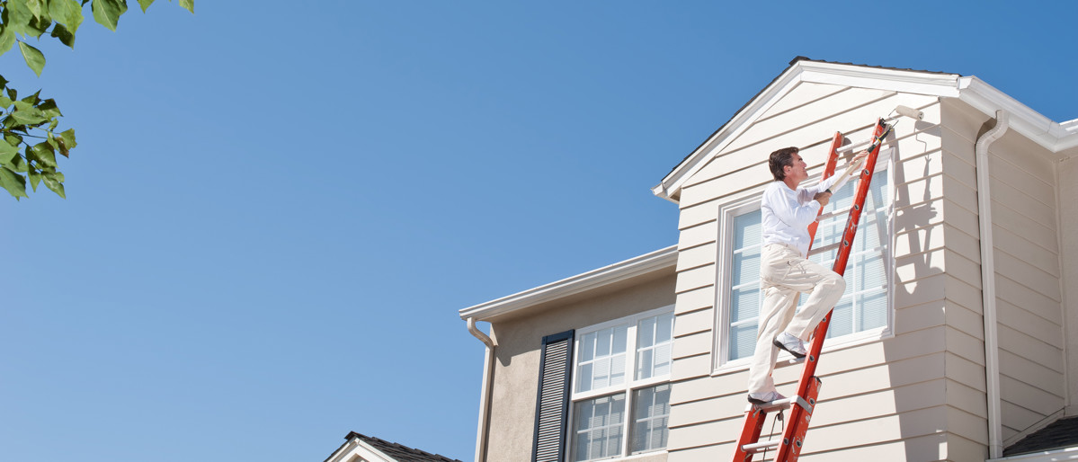 Husmålare i vita kläder målar fönsterkarm med roller på andra våningen av vitt hus i Cape Cod-stil